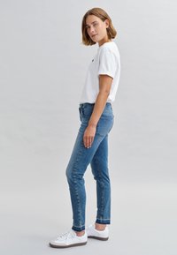 T-shirt à manches retroussées blanche associé à un jean skinny bleu clair et des baskets blanches avec des semelles marron, présentés sur un fond neutre.