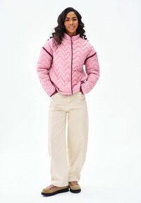 Žena s kučeravými vlasmi, oblečená v ružovej prešívanej bunde, béžových širokých nohaviciach a hnedých turistických topánkach, stojaca s rukami v vreckách.