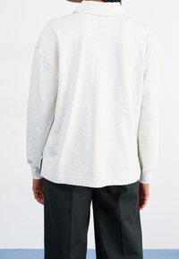 Ljust grå långärmad polo-skorta med en något lös passform, med en platt krage och sidoklyftor vid fållen. Slät textur.