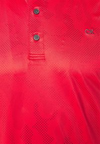 Červené tričko polo s tečkovaným vzorem, dvěma černými knoflíky a malým šedým logem "CK" na hrudní části. Měkká textura.