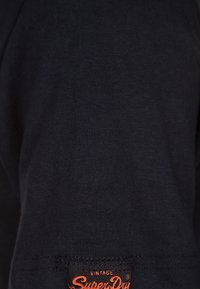 Černé bavlněné tričko s hladkou texturou. Má na lemu oranžovou značkovou etiketu "Vintage Superdry".