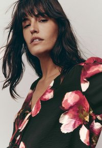 Camicia nera con grandi motivi floreali rosa e bianchi, design con scollatura a V e tessuto morbido e testurizzato. Maniche corte a sbuffo.
