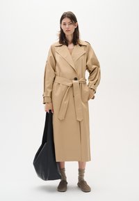Beige trench coat with a belted waist, large lapels, and structured sleeves. Model holds a black tote bag, wearing knit socks and brown shoes.
