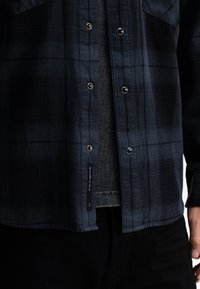 Geren check navy shirt met metalen drukkers, voorzien van een zak en een label met tekst. G gedragen over een grijze shirt, geaccentueerd door zwarte broek.