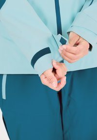 Lysblå jakkeærme med en mørkeblå velcro-detalje. Materialet fremstår glat og viser et struktureret design samt funktionelle manchetter.