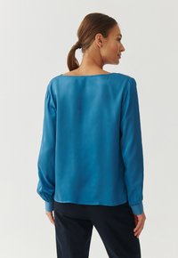 Blusa di raso blu con maniche lunghe, scollatura ampia e polsini arricciati. Il tessuto ha una superficie liscia e una leggera lucentezza.
