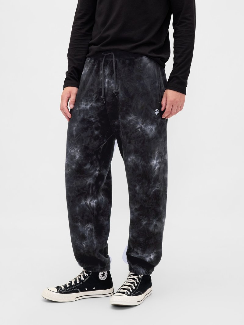 Person in schwarz-grauem Batik-Jogginghose mit elastischen Bündchen, schwarzem Langarmshirt und schwarzen High-Top-Sneakern vor einem weißen Hintergrund.