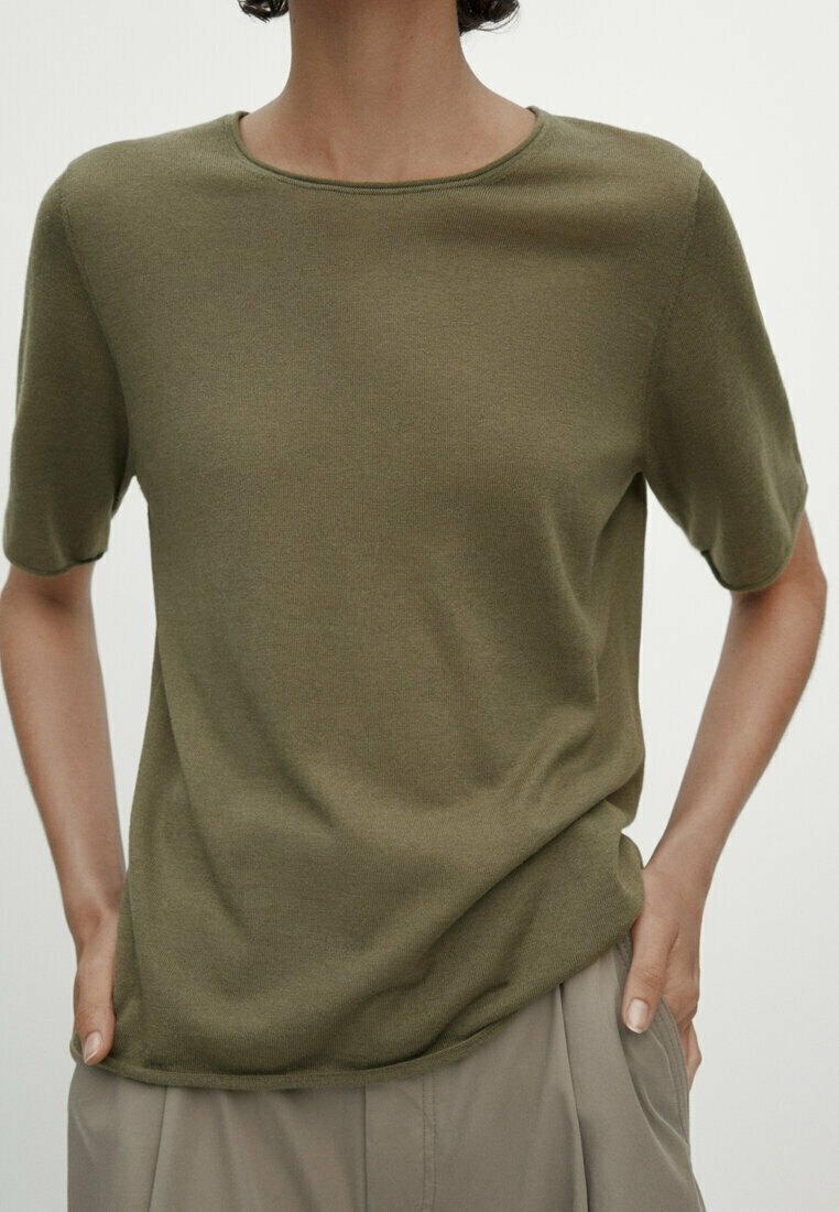 Personne portant un haut tricoté manches courtes vert olive et un pantalon beige, les mains dans les poches sur un fond uni.