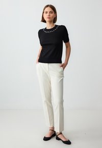 Black short-sleeve top with embellishments at the neckline, paired with cream trousers and black flats. Smooth fabric with a fitted design.