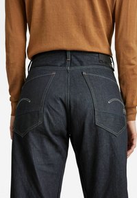 Jeans in denim blu scuro con vita alta, tasche posteriori con alette inclinate e dettagli di cucitura a contrasto. L'outfit include una maglietta marrone.