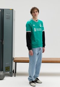 Grønn Liverpool fotballdrakt med svarte ermer, hvite striper og logo. Brukt med lys blå jeans og svart-hvite joggesko.