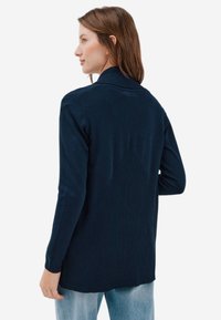 Cardigan tricot marine avec texture côtelée au dos, manches longues et coupe décontractée. Design simple, sans fermetures ni motifs visibles.