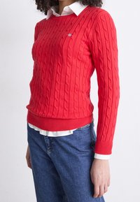 Femme portant un pull rouge en maille torsadée par-dessus une chemise blanche à col, associée à un jean bleu, debout devant un fond uni.