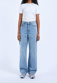 Světle modré široké džíny s vysokým pasem, pět kapes, klasická denimo látka, v kombinaci s přiléhavým bílým tričkem a šedými teniskami.