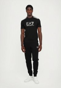 Svart t-shirt med EA7-märke och grå axeldetaljer samt svarta mjukisbyxor. Modellen bär vita sneakers och står mot en enfärgad bakgrund.