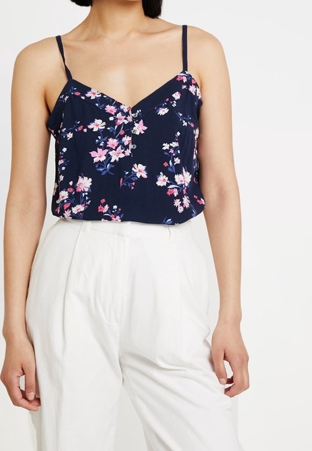 Femme portant un haut sans manches bleu marine à fleurs avec fines bretelles et un pantalon blanc taille haute, debout devant un fond blanc uni.