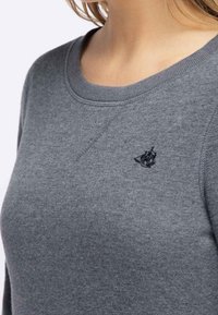 Sudadera gris con cuello redondo, que presenta un pequeño logo bordado en el pecho. Textura suave, con puños y dobladillo acanalados.
