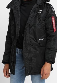 Veste matelassée noire avec un col montant, fermeture éclair et boutons, plusieurs poches, poignets côtelés et détail d'étiquette en tissu rouge sur la manche.