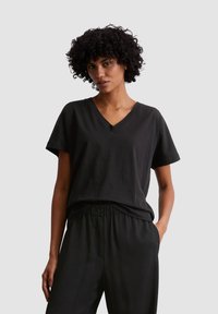 Zwarte V-hals t-shirt gemaakt van zachte katoen. Korte mouwen, relaxte pasvorm. Draagt bijpassende zwarte broek, beiden weergegeven tegen een neutrale achtergrond.