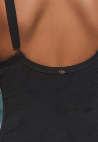 Gros plan sur le haut du dos d'une personne portant un maillot de bain noir texturé avec de fines bretelles réglables et une petite boucle décorative dorée.