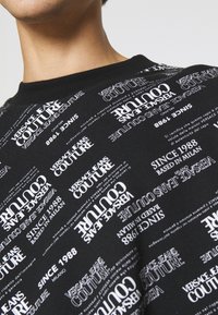 Černá mikina s bílým potiskem, který zahrnuje text "Versace Jeans Couture" a "Since 1988" v různých velikostech písma a orientacích.