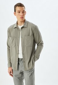 Ljusgrå vävd skjortjacka med två framfickor, tryckknappar, långa ärmar och en avslappnad passform, lager på lager över en vit skjorta.