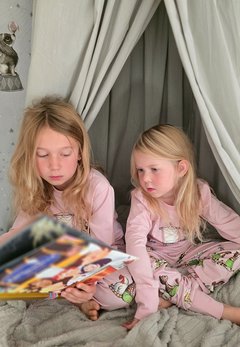 Two children in pink pajamas with cartoon patterns sit on a gray blanket, reading a colorful book inside a fabric tent.