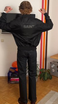 Melna bumbieru jaka ar uzrakstu "SAINTS" aizmugurē, apvienota ar melnām biksēm. Iestatījums ar koka grīdu un krāsainiem sienu akcentiem.