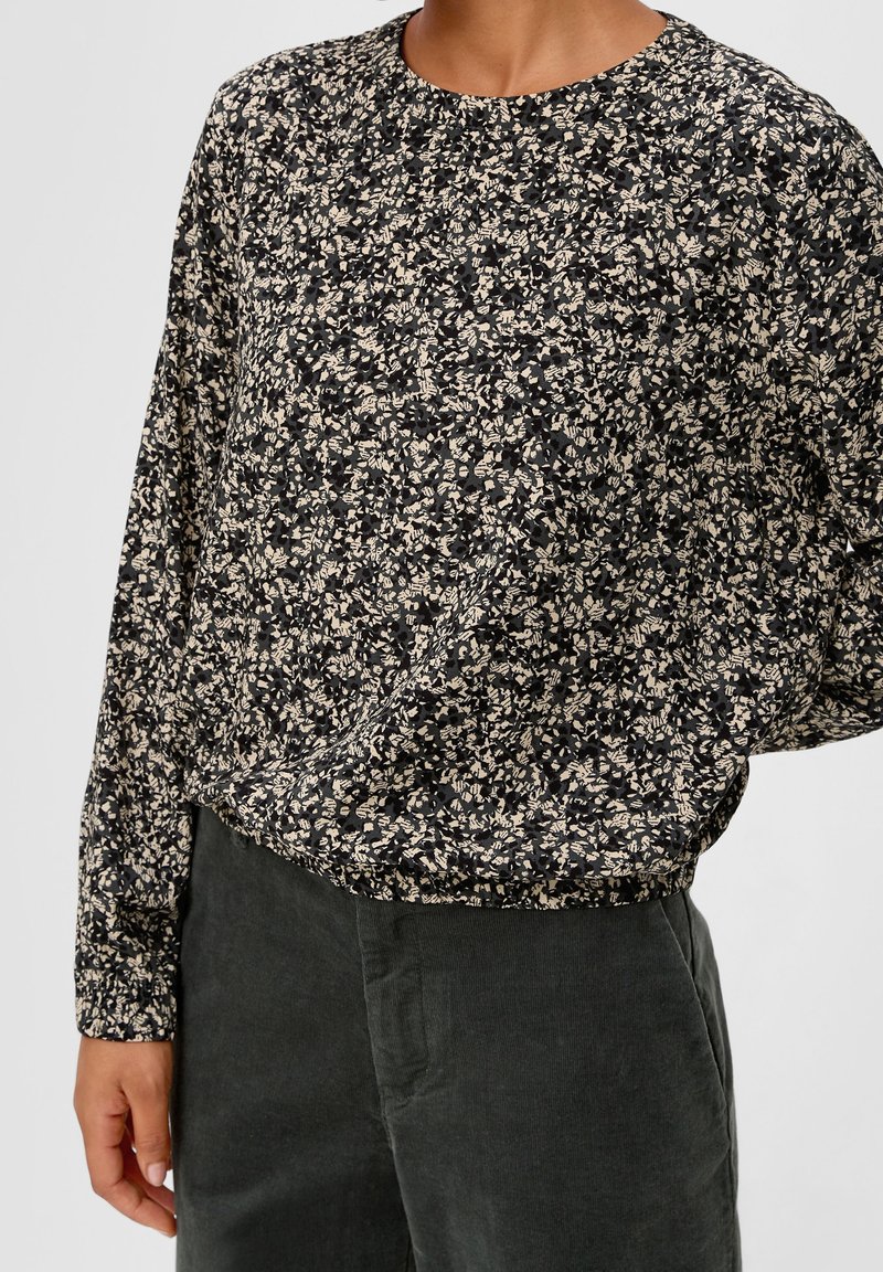 Blouse fleurie noire et blanche à manches longues associée à un pantalon en velours côtelé vert foncé portée par une personne sur un fond neutre.