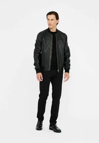 Giacca bomber nera in pelle con cerniera frontale, collo a coste e polsini aderenti, abbinata a pantaloni neri e scarpe lucide.