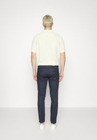 Man med kort blont hår iförd en kortärmad krämfärgad skjorta, mörka slim-fit byxor och vita sneakers som står vänd mot en enfärgad vit vägg.