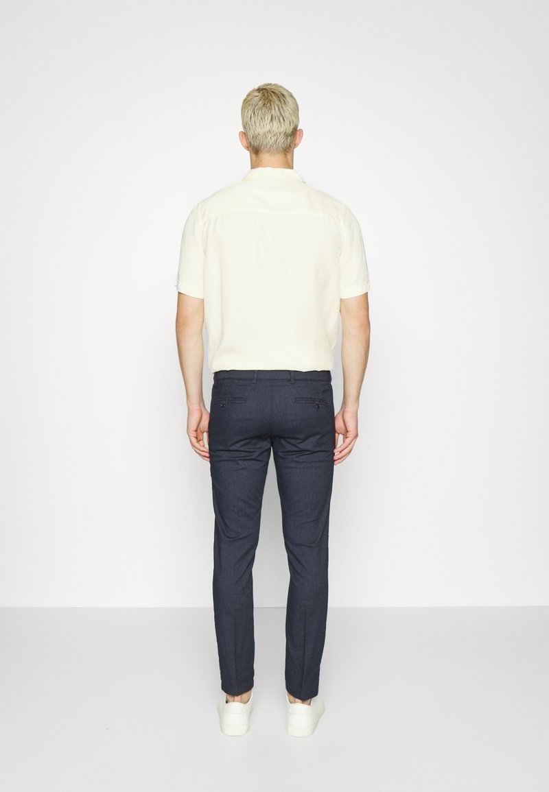 Man med kort blont hår iförd en kortärmad krämfärgad skjorta, mörka slim-fit byxor och vita sneakers som står vänd mot en enfärgad vit vägg.