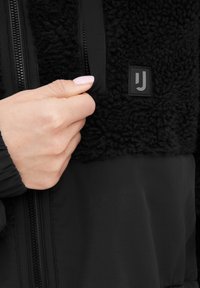 Giacca nera con un materiale superiore morbido e peloso, sezione inferiore liscia e zip. Un logo notevole arricchisce il design.