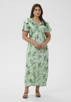 Žena u svjetlozelenoj haljini na cvjetni uzorak s volanima na rukavima i crnim sandalama s otvorenim prstima, stoji na jednobojnoj bijeloj pozadini.