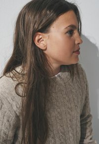 Jasnobeżowy sweter z warkoczem o okrągłym dekolcie, noszony na białej bluzce. Ciemnobrązowe, falowane włosy częściowo zasłaniają ramię.