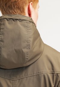 Olivově zelená bunda s kapucí a hladkou matnou texturou. Má tvarovanou kapuci a viditelné švy podél švů.