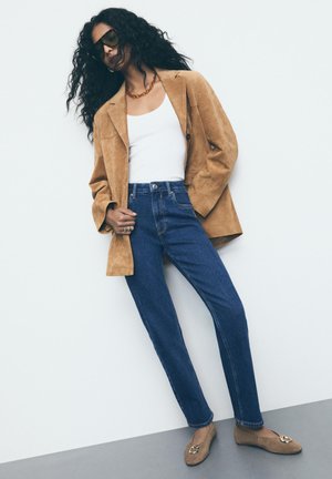 Blazer in suede tan sopra un top bianco, abbinato a jeans a vita alta blu e ballerine marroni con accessori decorativi. Indossati occhiali da sole trasparenti.