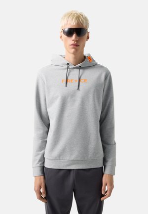 Jong persoon met kort blond haar draagt donkere zonnebril en een lichtgrijze hoodie met oranje "FIRE+ICE" tekst, gecombineerd met donkere broek.