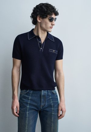 Άνδρας με κατσαρά μαλλιά που φοράει μαύρα γυαλιά ηλίου, μπλε πουλόβερ πόλο με λευκή ραφή και μπλε τζιν, κοιτάζοντας πλάγια.