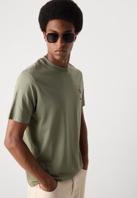Olijfgroene katoen t-shirt met ronde hals, voorzien van een klein geborduurd ontwerp op de borst. Model draagt een zonnebril.