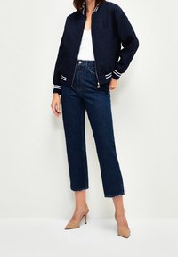 Granatowa kurtka typu bomber z fakturowanej tkaniny, z białymi paskowanymi mankietami i kołnierzem, w zestawie z wysokim stanem dżinsowych spodni i cielistymi szpiczastymi szpilkami.