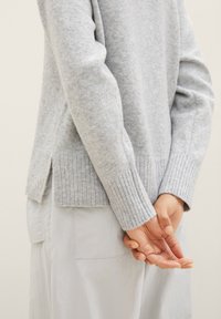 Pull gris clair avec ourlet et poignets côtelés, fabriqué en tissu doux. Présente des fentes latérales pour le confort, associé à un pantalon gris clair.