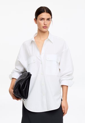 Femme portant une chemise blanche oversize à manches retroussées, tenant un sac à main en cuir noir sur un fond blanc uni.