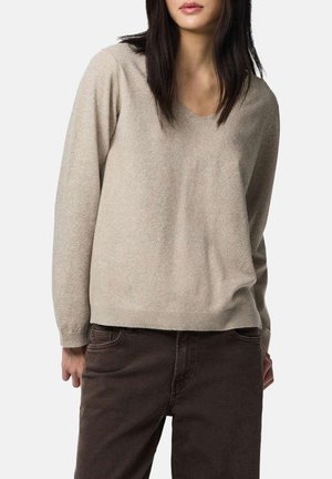 Mujer con suéter beige de manga larga y cuello en V y pantalones marrón oscuro, de pie con las manos relajadas a los lados contra un fondo blanco.