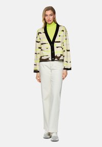 Strickjacke mit schwarzem V-Ausschnitt, die unregelmäßige grüne, braune und beige Muster aufweist. Getragen über einem leuchtend grünen Rollkragenpullover und weißen Hosen.