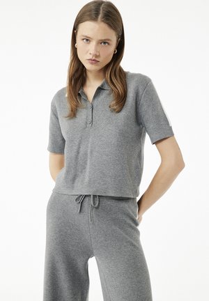 Joven mujer con cabello liso y castaño, llevando una camisa polo gris de manga corta y pantalones a juego con cordón, sobre un fondo blanco.