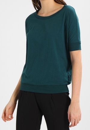 Kvinde iført en teal kortærmet top med rund halsudskæring og sorte bukser, stående mod en helt hvid baggrund.