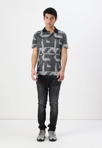 Camisa polo con patrón geométrico en blanco y negro, con cuello negro, combinada con jeans oscuros y zapatillas deportivas grises.