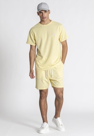 Junger Mann trägt ein blassgelbes T-Shirt und passende Shorts, weiße Sneakers und eine hellgraue Baseballkappe, steht vor einem einfarbigen Hintergrund.