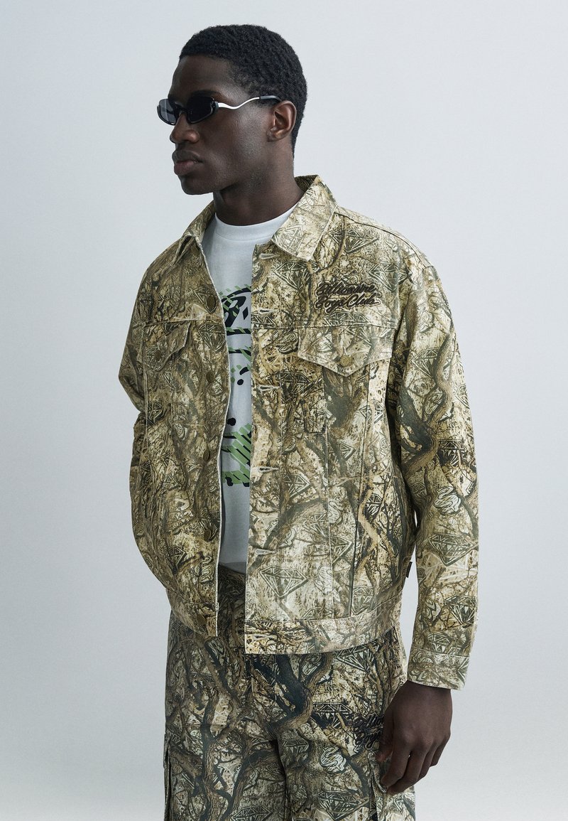 Μπουφάν και παντελόνι με καμουφλάζ σε αποχρώσεις μπεζ και πράσινου, κατασκευασμένα από ανθεκτικό ύφασμα. Διαθέτουν τσέπες στο στήθος και από μέσα υπάρχει ένα γραφικό λευκό μπλουζάκι.